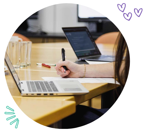A person taking notes at a conference table with laptops, glasses of water, and a pen, in a meeting room.