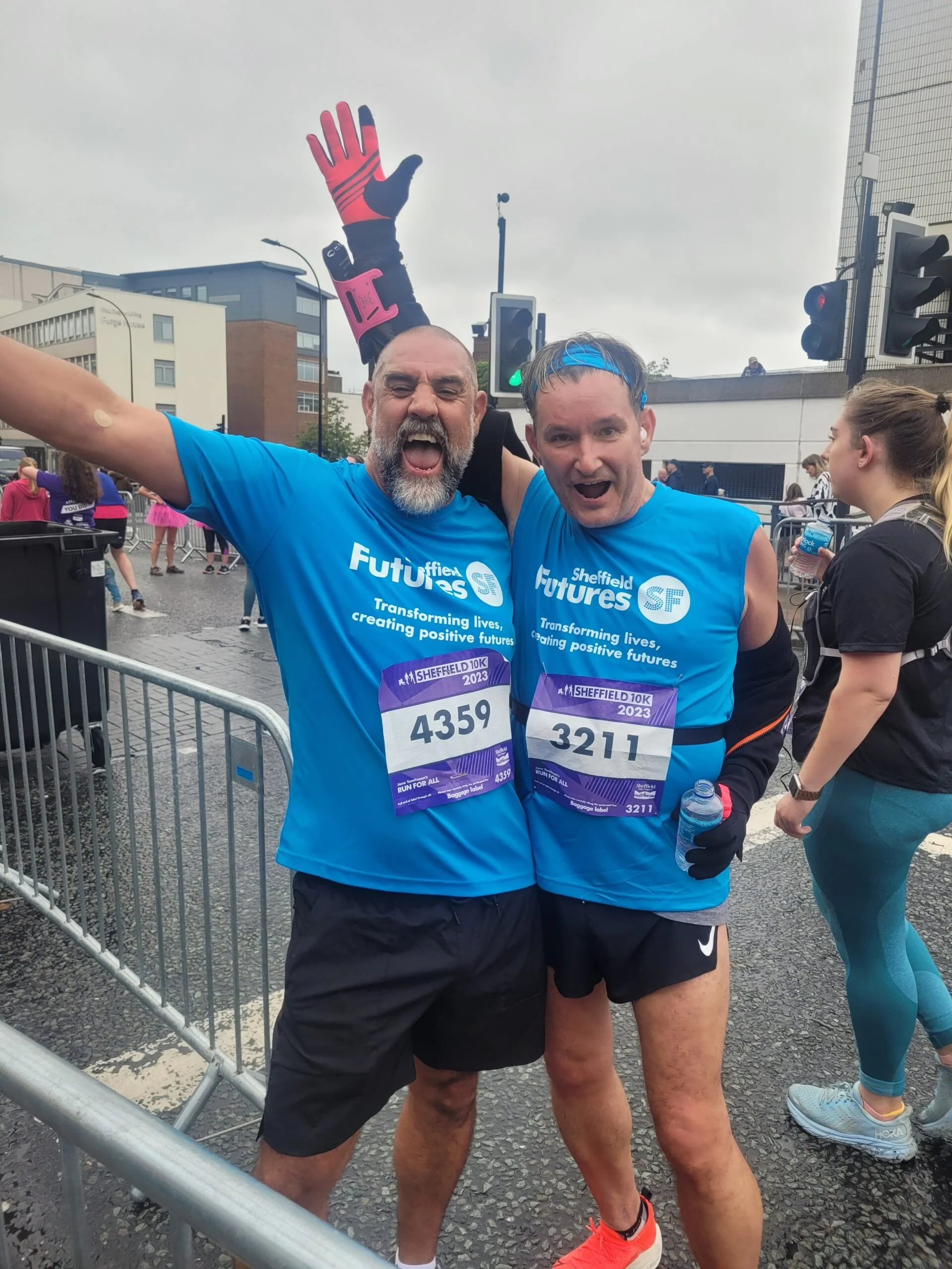 Two men in blue shirts celebrating at a marathon event, with one raising his arm and both smiling, wearing race bibs numbered 4359 and 3211.