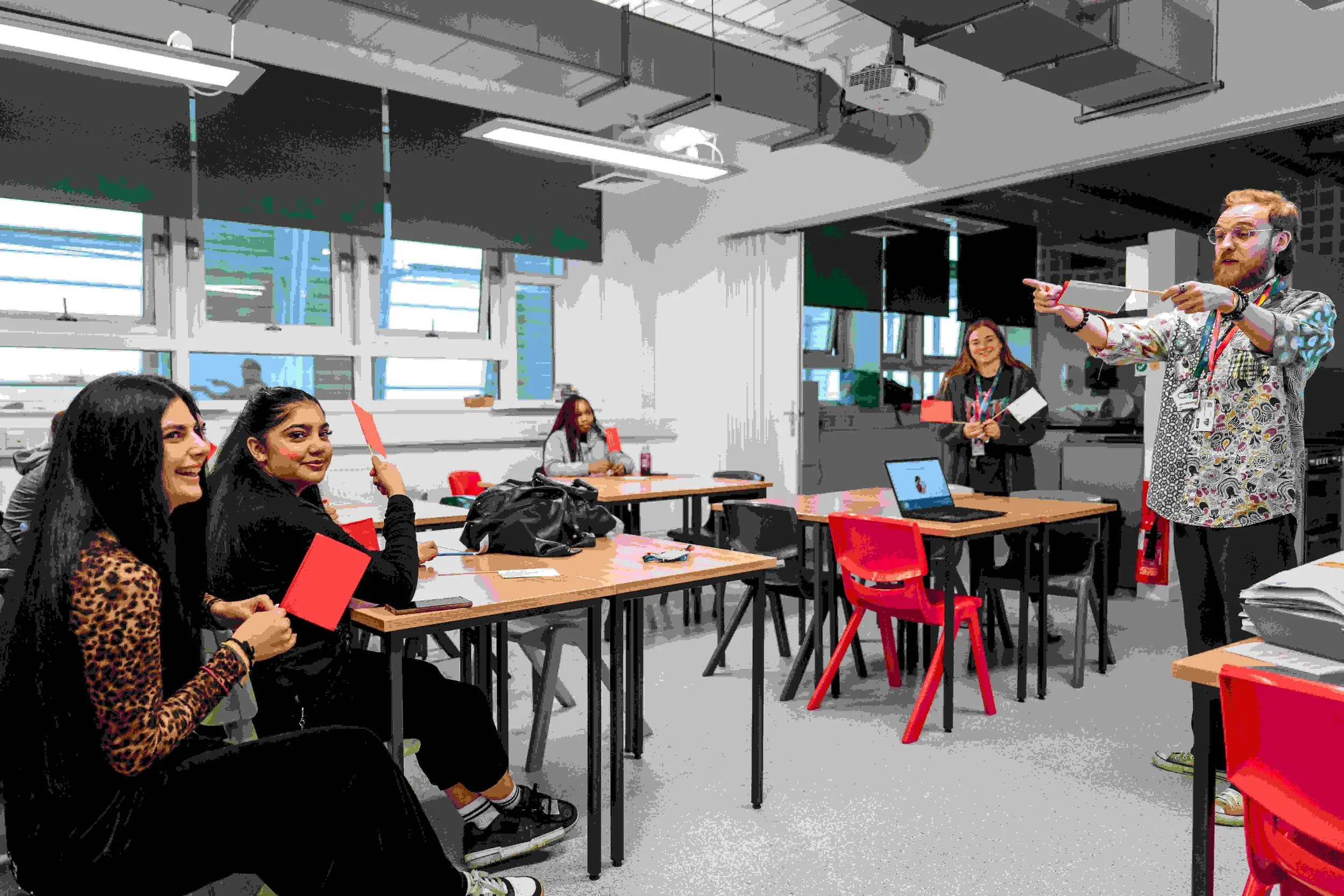 A classroom with students sitting at desks, some holding red notebooks, and a teacher standing at the front pointing. The room has large windows, a projector, and a laptop on a desk.