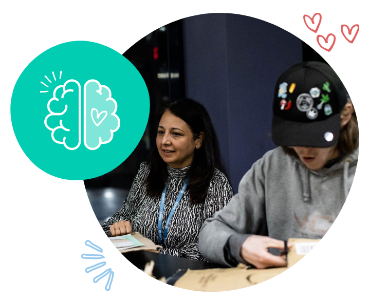 Two women sitting at a table, one appears to be explaining something while the other looks bored. Decorative graphics include a brain with a heart inside, small hearts, and a few lines.