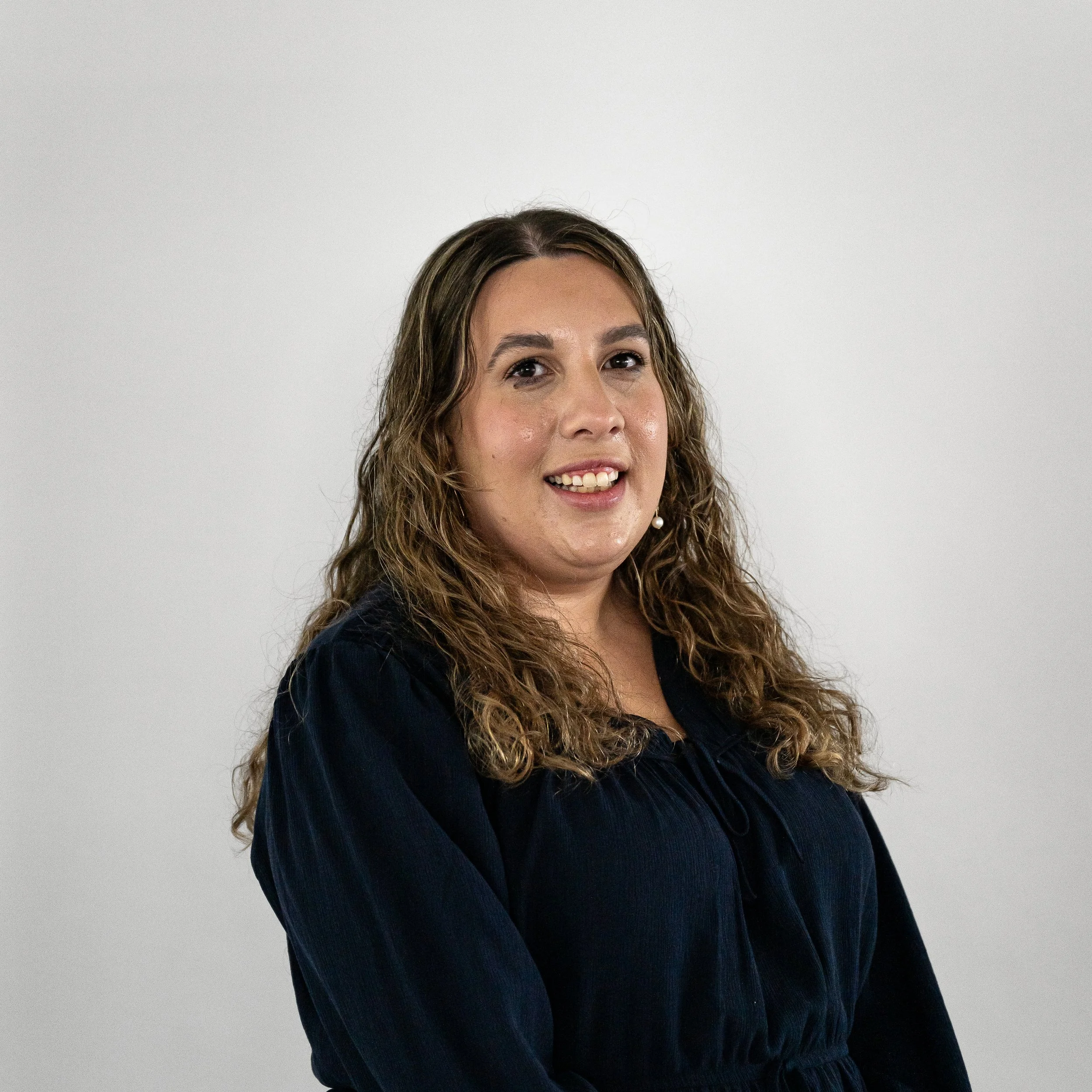 A woman with curly, shoulder-length hair, wearing a dark blue blouse, smiling at the camera against a plain light gray background.
