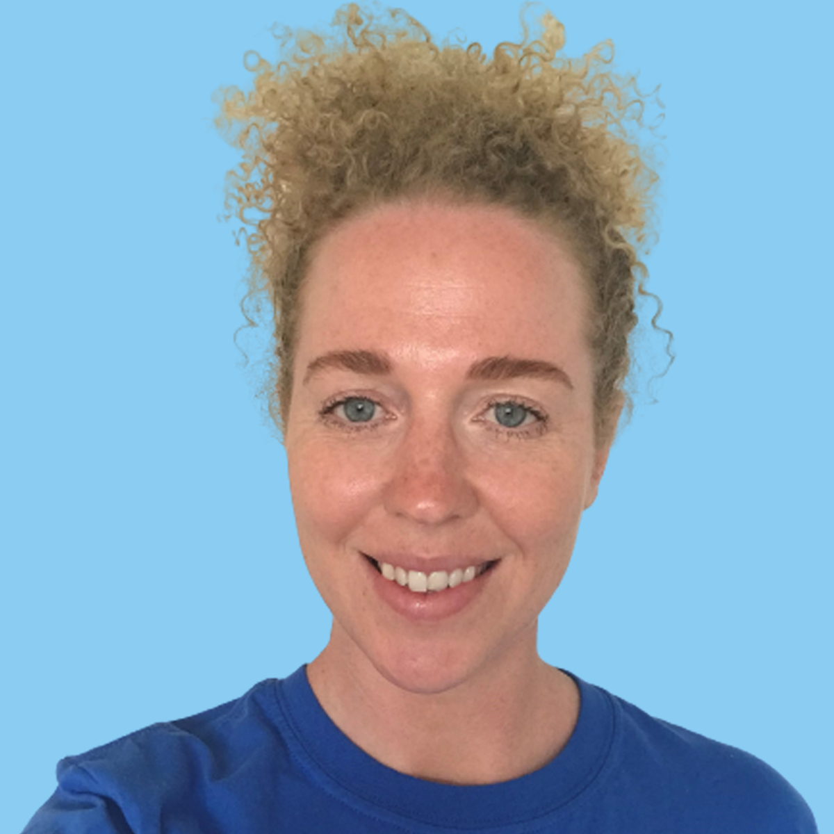 A woman with curly blonde hair, light skin, and blue eyes smiling, wearing a blue shirt, standing against a light blue background.