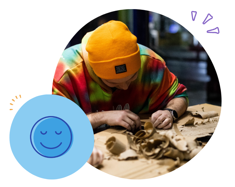 Person wearing a yellow beanie and rainbow-colored hoodie working on a craft project at a wooden table, with paper and crafting materials around.