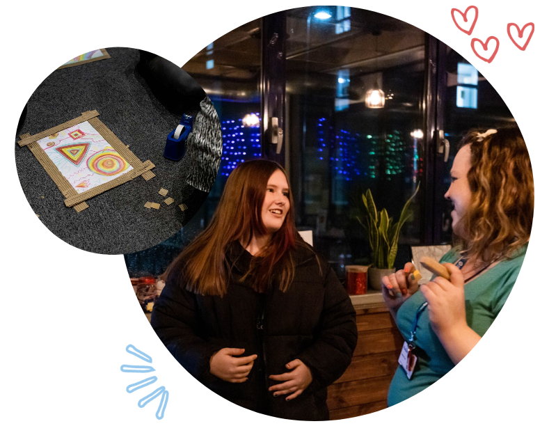 Two women engaged in a conversation indoors at night, with a large window reflecting colorful lights. One woman has long red hair and is wearing a black jacket, and the other woman has curly hair and is wearing a turquoise shirt with an ID badge. There is a small potted plant on the windowsill.