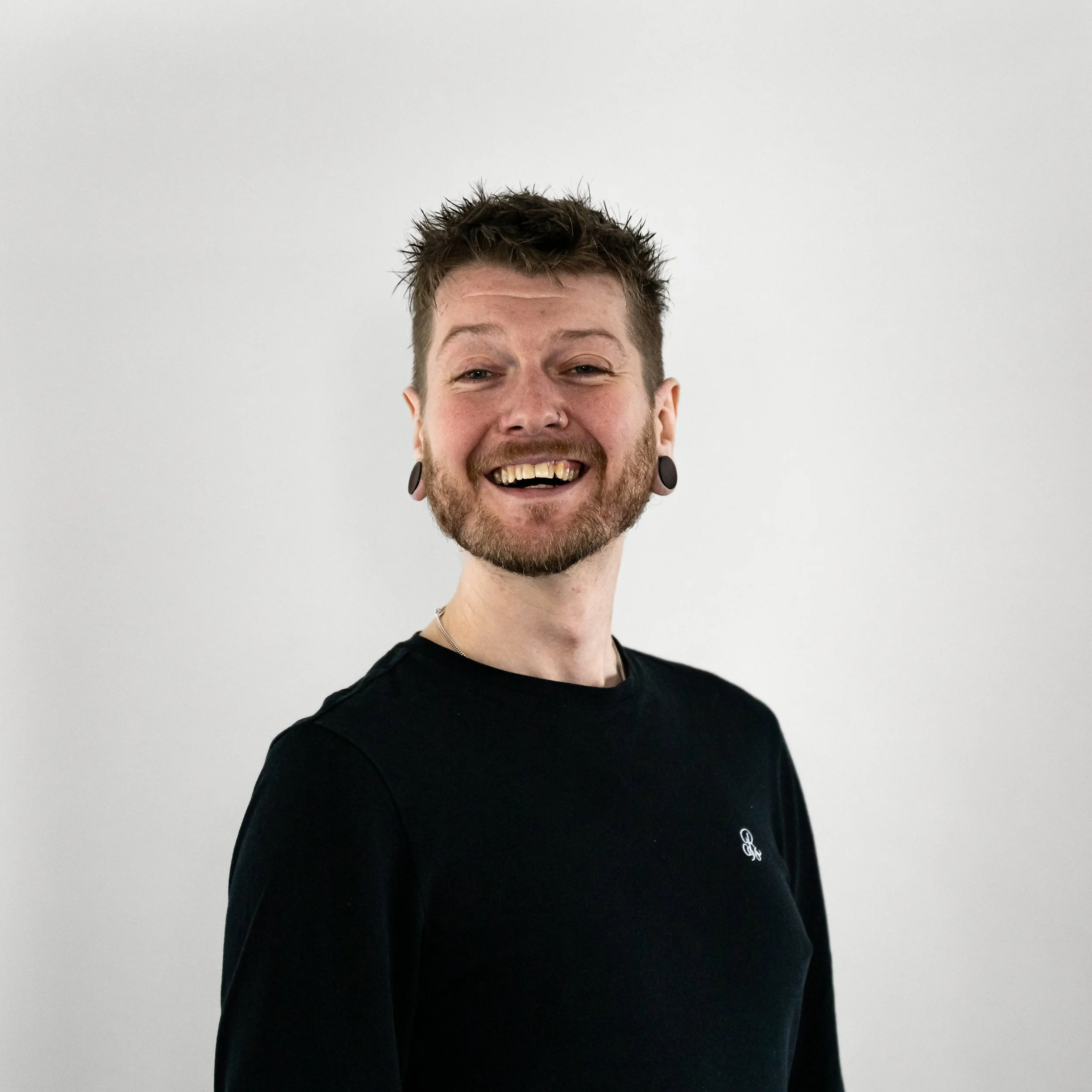 Portrait of a smiling man with tattoos, wearing a black shirt, standing against a plain white background.