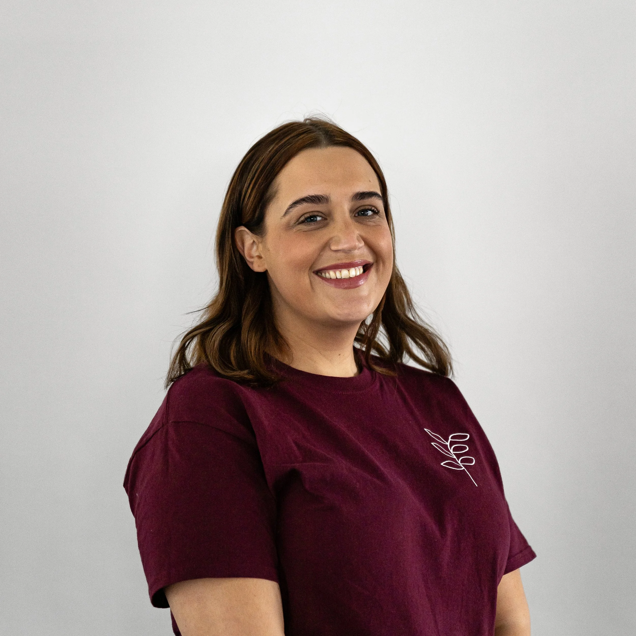 A young woman with shoulder-length brown hair smiling and wearing a maroon T-shirt with a white design on the chest, standing against a plain gray background.