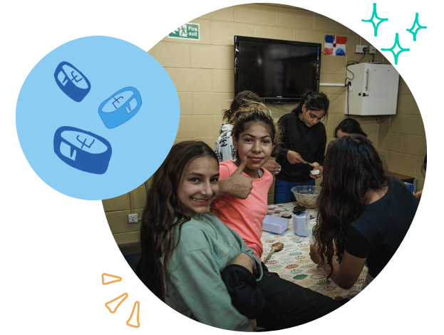 Group of girls sitting around a table in a room with beige walls, with a TV mounted on the wall and several objects on the table, engaged in an activity or craft.