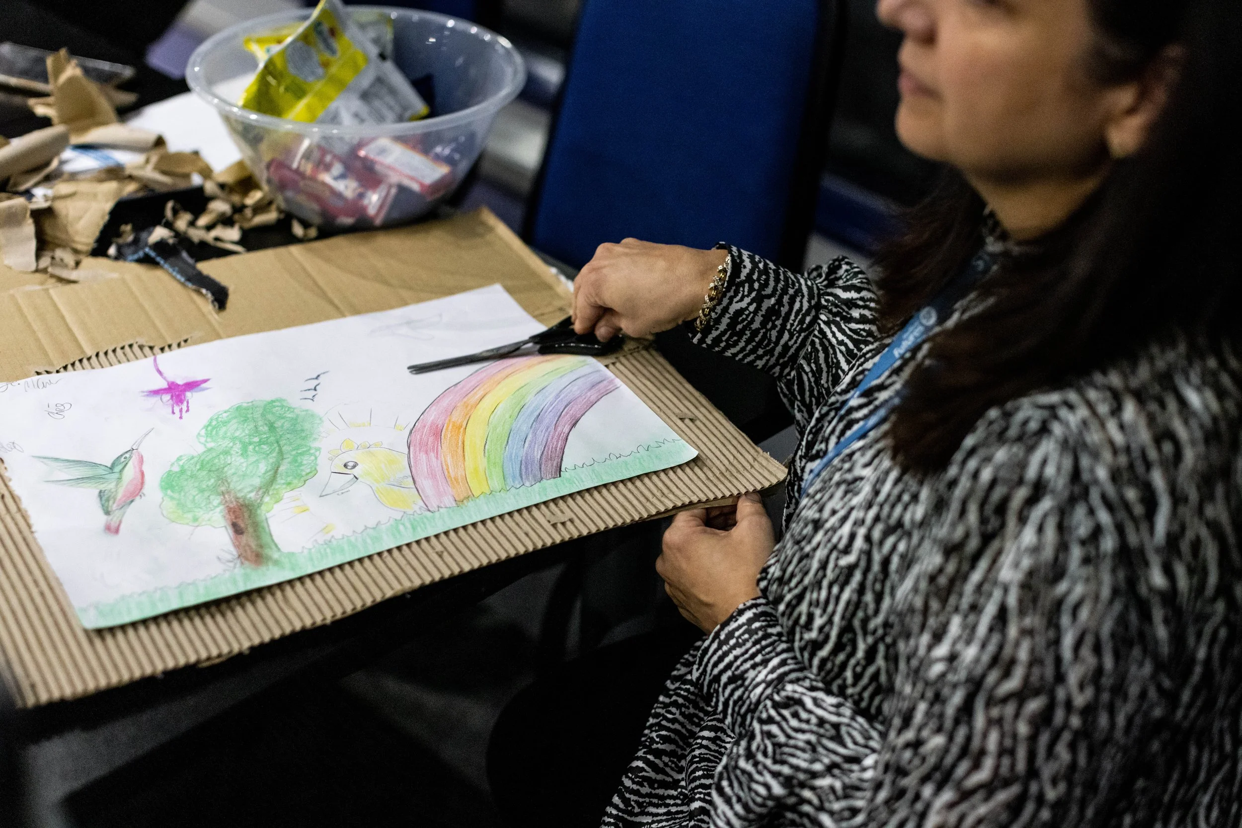 A woman coloring a drawing of a rainbow, a tree, a unicorn, and a bird on a piece of paper on a cardboard mat, with art supplies in a clear plastic bowl on a table.