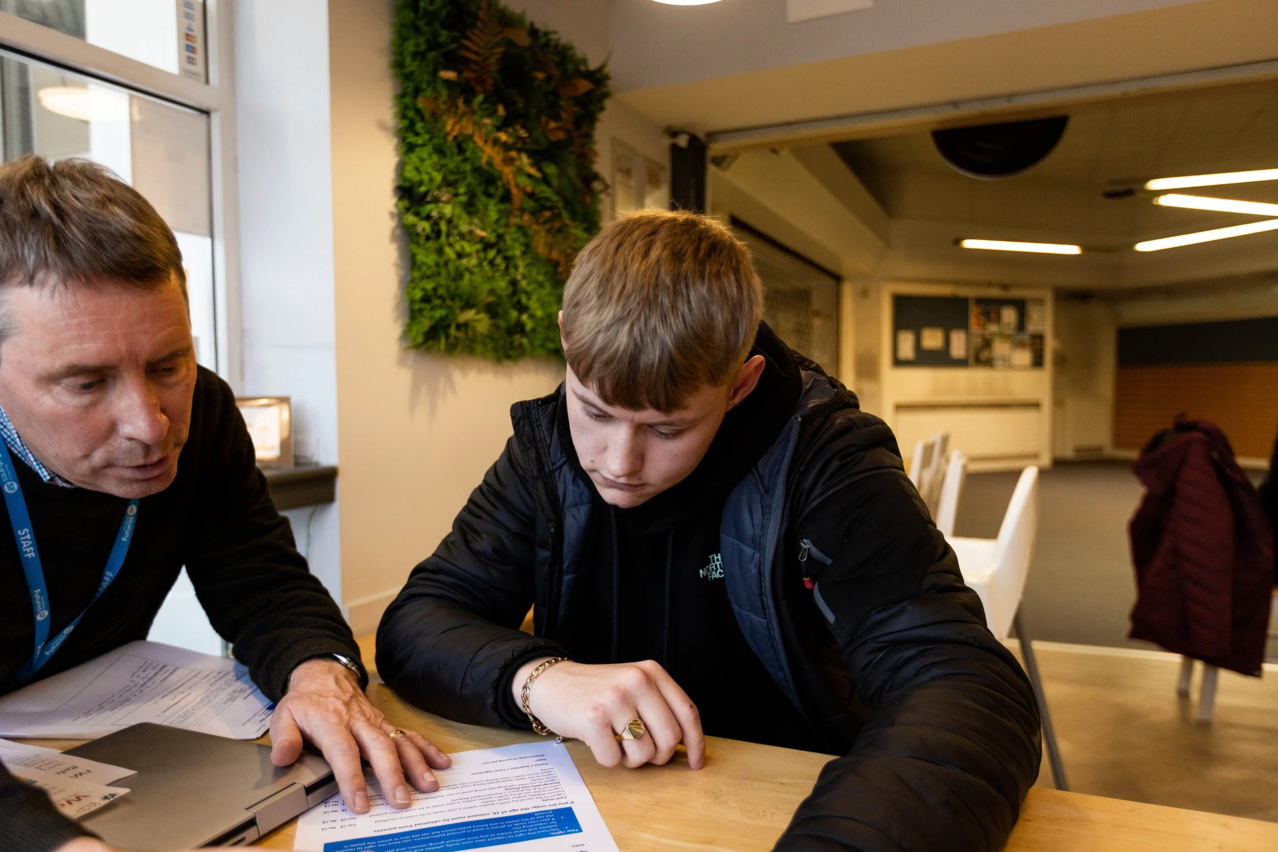 A young man and an older man sitting at a table looking at documents, with the young man pointing at the papers, in a well-lit indoor space with a green wall decoration in the background.