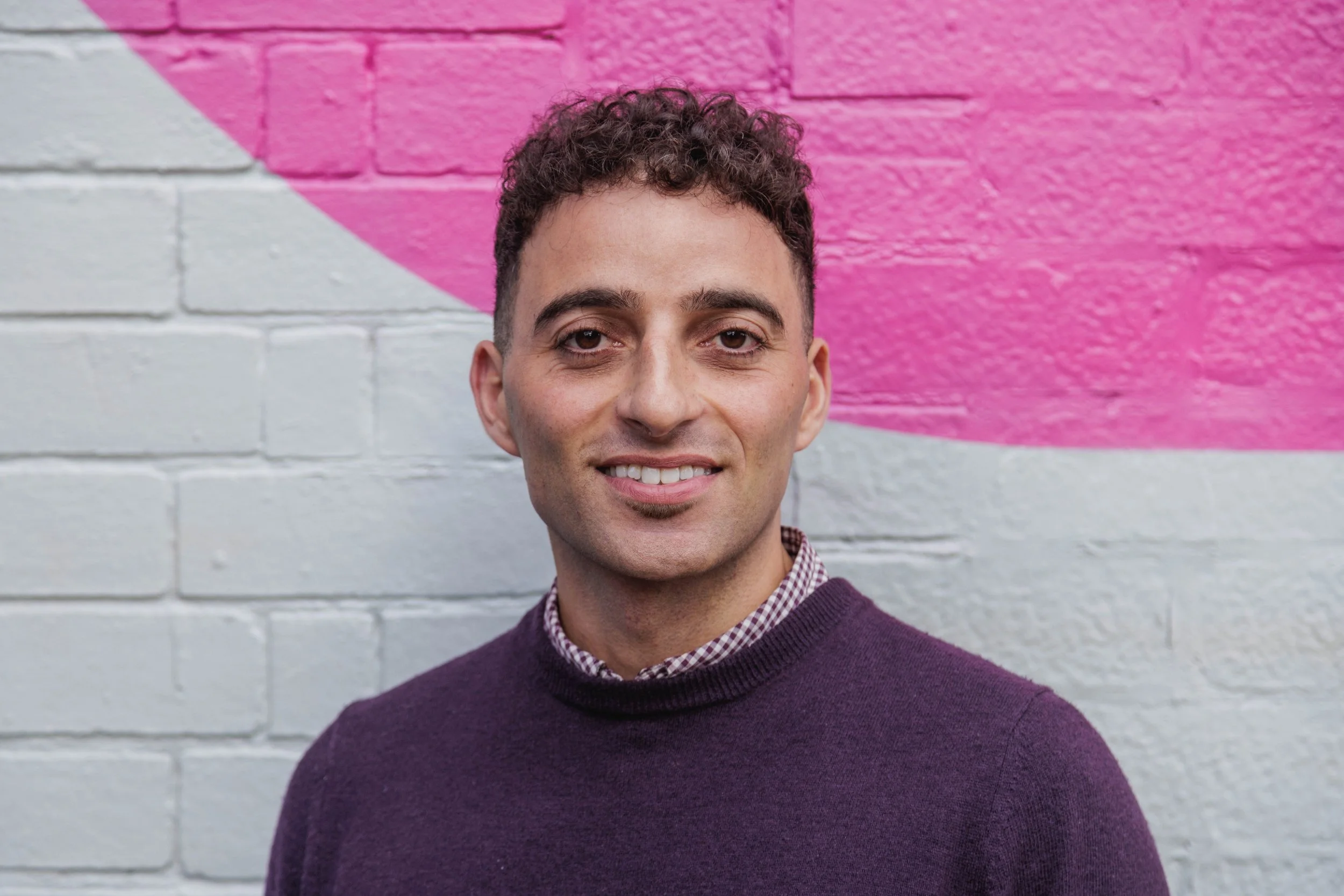 A young man with curly brown hair, wearing a purple sweater over a checked shirt, standing against a white brick wall with pink graffiti.
