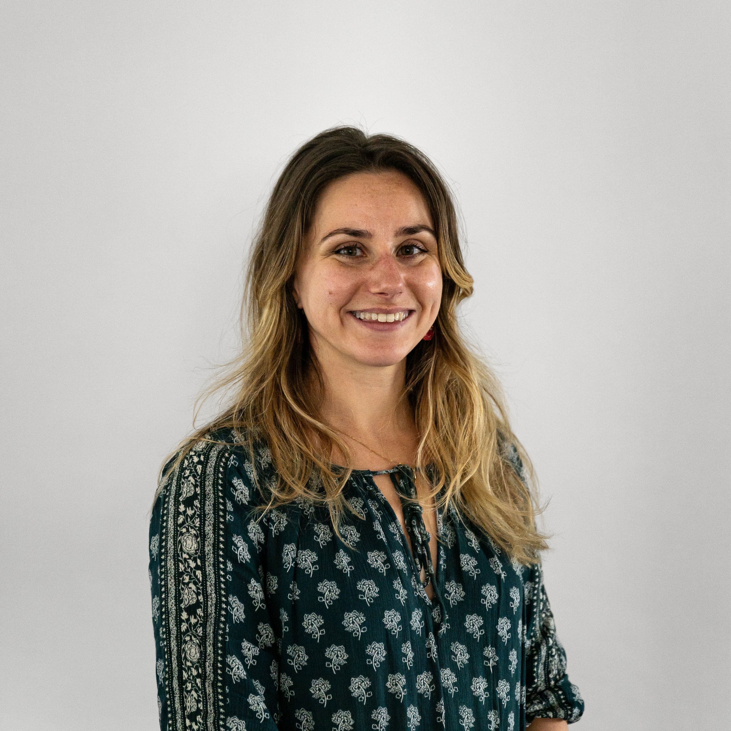 A smiling woman with shoulder-length wavy hair wearing a dark patterned blouse against a plain light gray background.