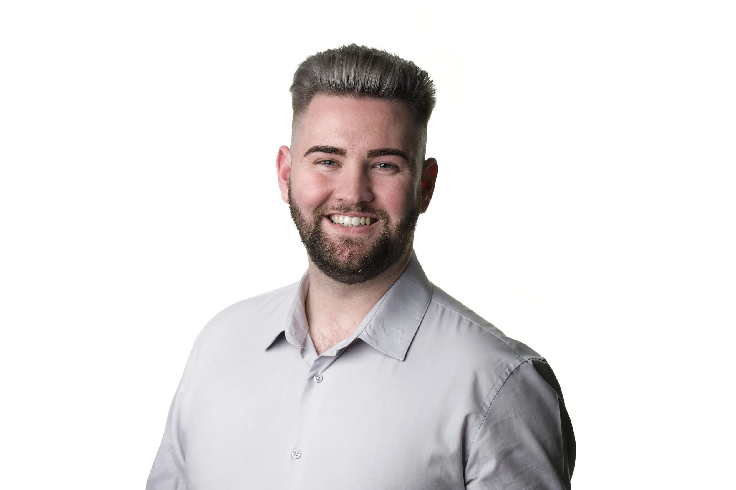Portrait of a smiling young man with a beard, short brown hair, and wearing a light gray button-up shirt, against a white background.