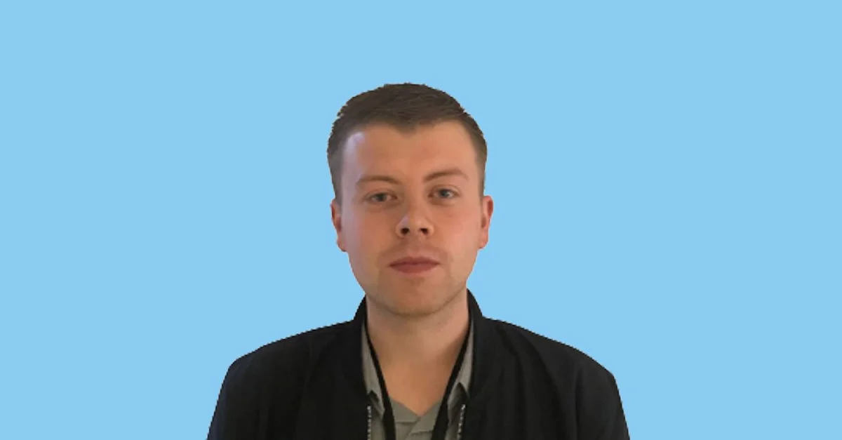 A young man with short brown hair, wearing a black jacket over a gray shirt, stands against a solid light blue background.