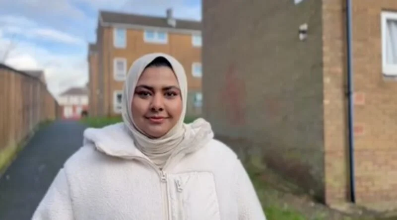 A woman wearing a cream-colored hijab and a white fleece jacket standing outdoors in a residential area with brick buildings and a paved alleyway.