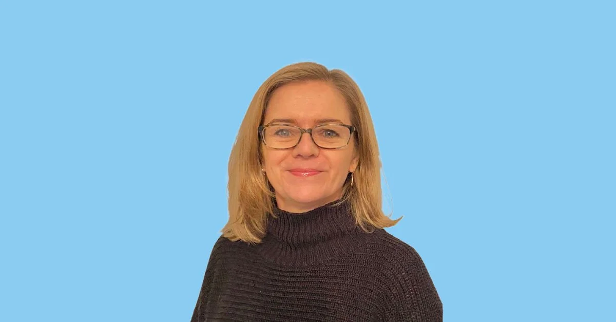 A woman with shoulder-length blonde hair, glasses, and a black turtleneck sweater, smiling against a light blue background.