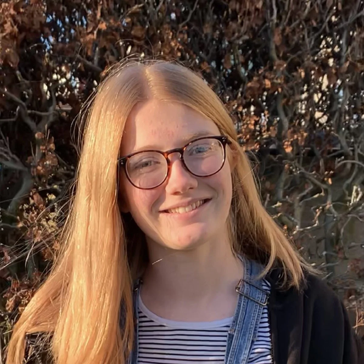 A young woman with long red hair, glasses, and a striped shirt smiling outdoors with brown shrubbery in the background.