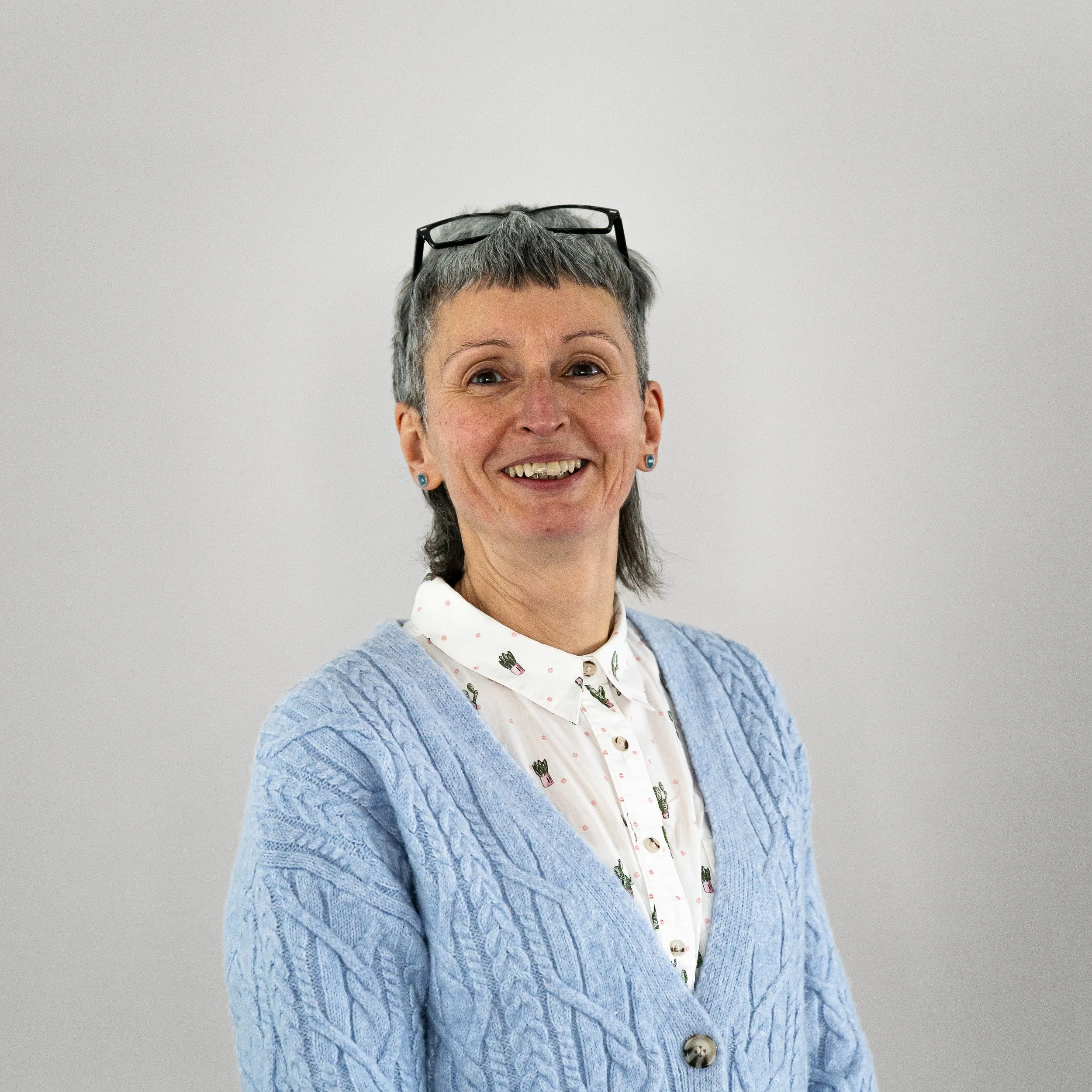 A smiling middle-aged woman with short gray hair and glasses resting on her head, wearing a light blue cable-knit cardigan over a white blouse with a small plant pattern, standing against a plain gray background.