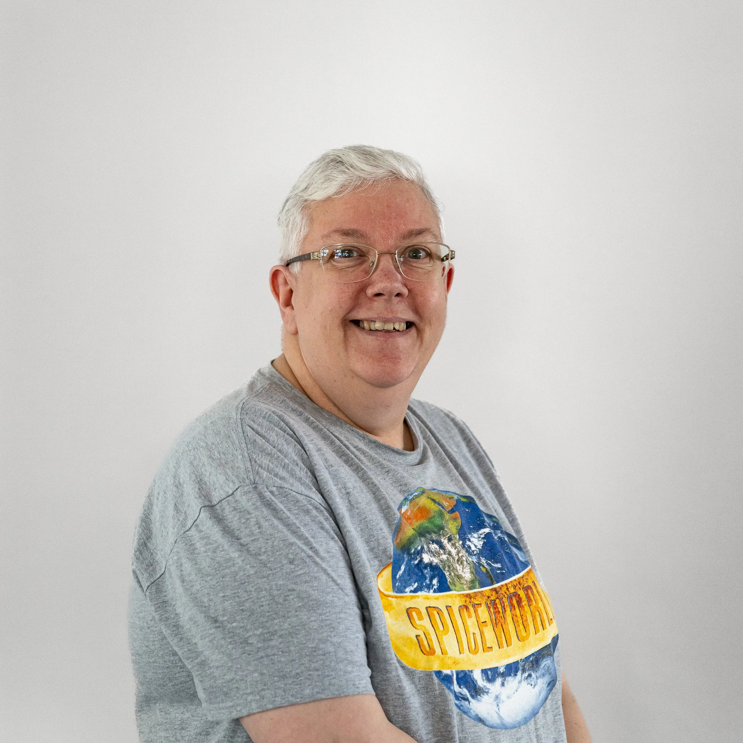A smiling man with glasses and gray hair, wearing a gray T-shirt with a globe graphic and 'SPICEWORLD' text, standing against a plain white background.