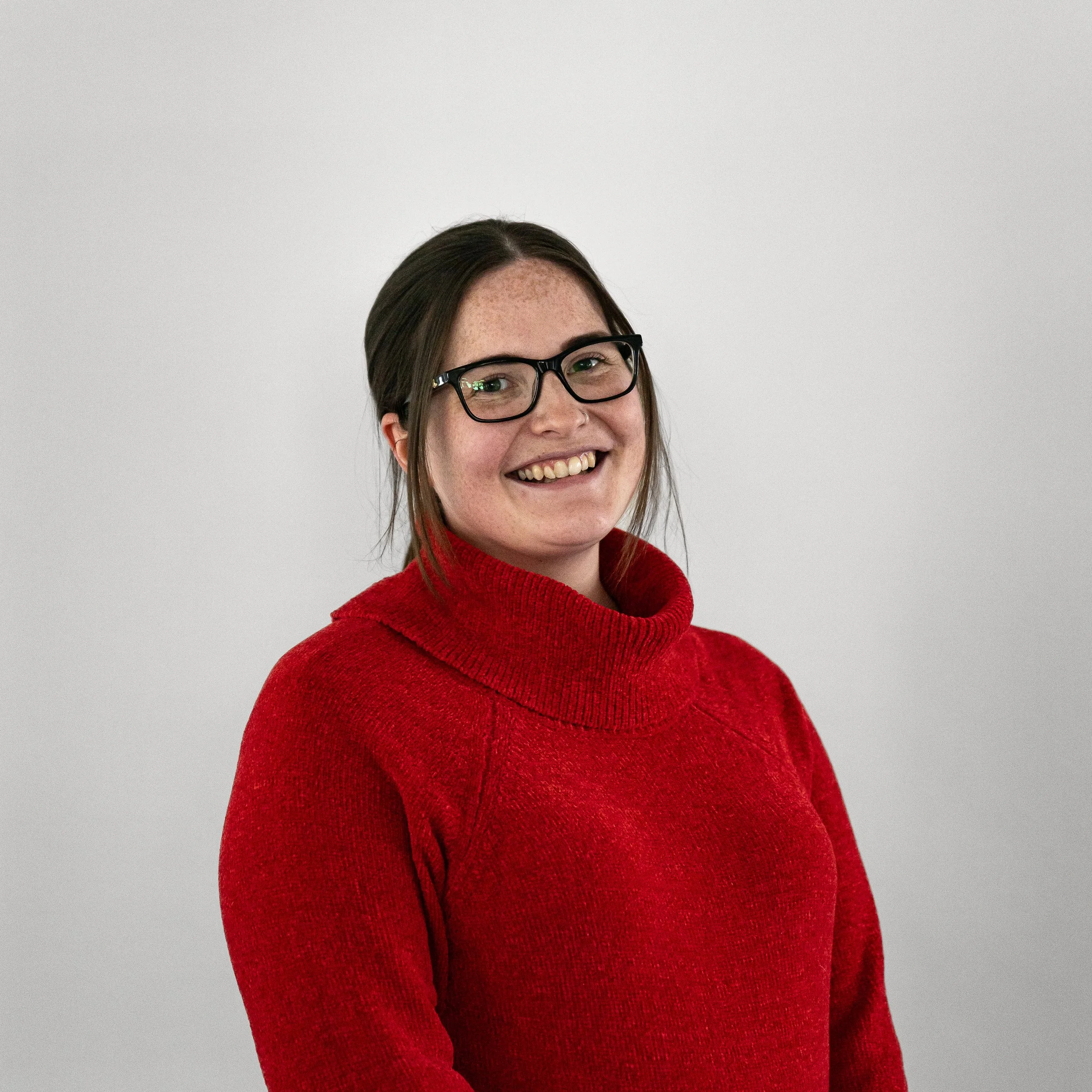 A woman with glasses and brown hair, smiling and wearing a red turtleneck sweater, standing against a plain white background.