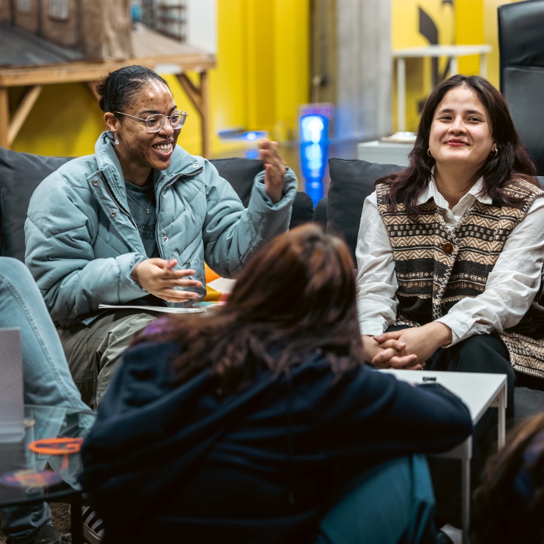 Two women sitting on a black couch, engaged in conversation in an indoor space with a yellow wall, shelves, and a water dispenser in the background.