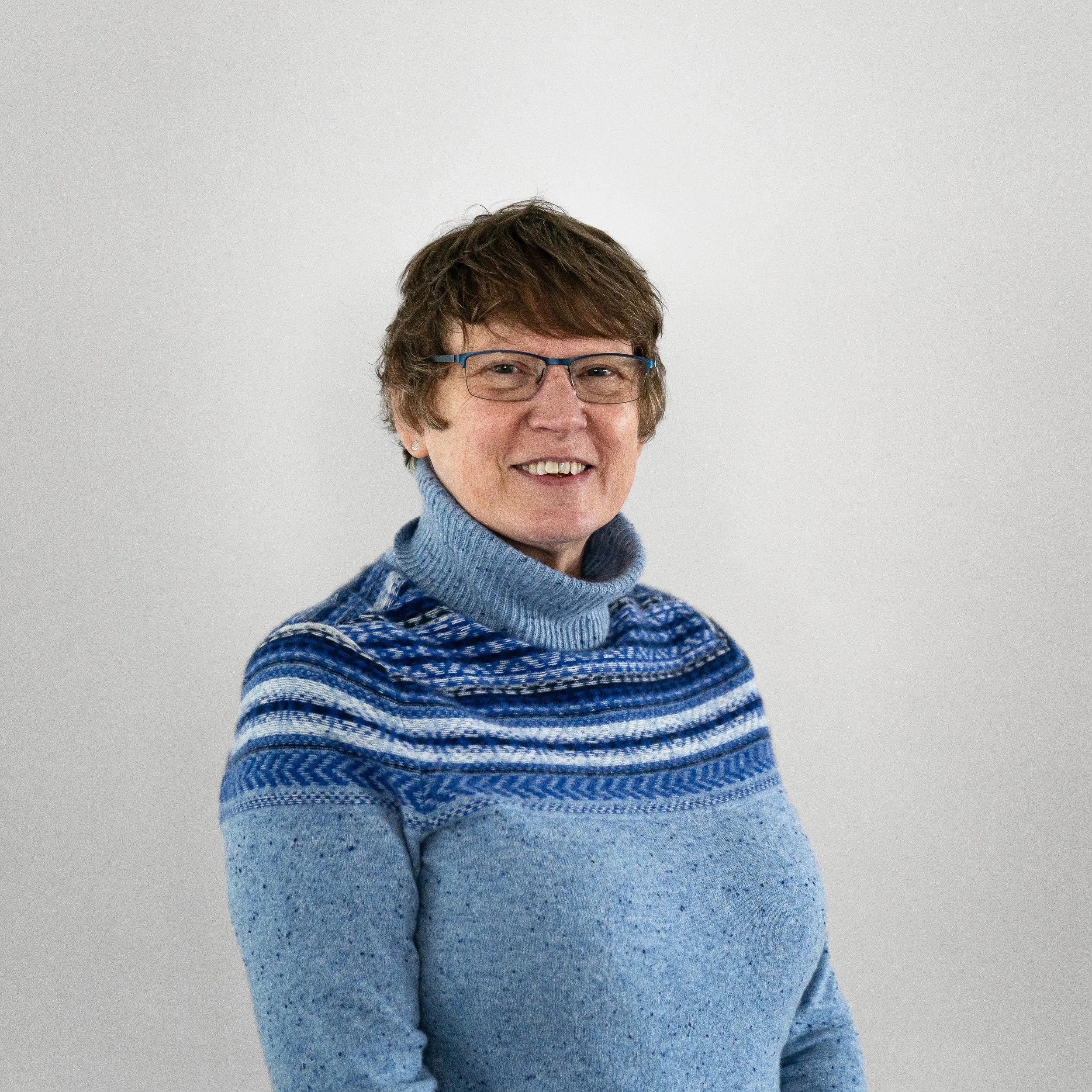 A woman with short brown hair, glasses, and a fair complexion smiling, wearing a light blue turtleneck sweater with a blue patterned yoke, standing against a plain light gray background.