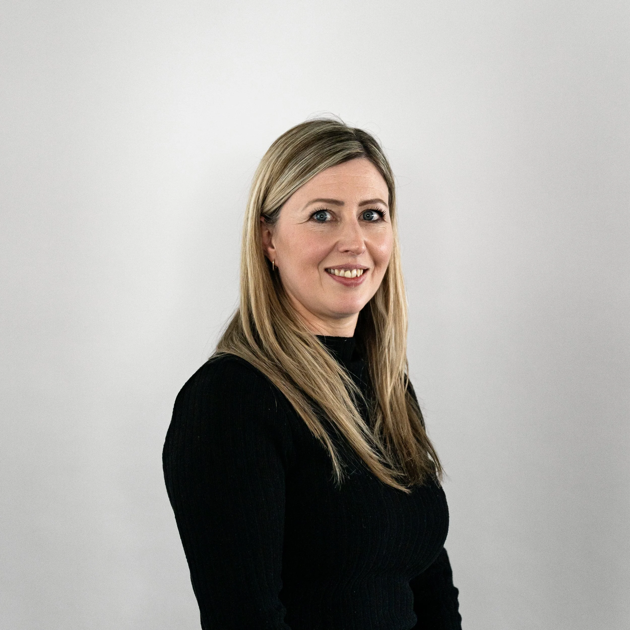 A woman with long blonde hair smiling at the camera, wearing a black top, standing against a plain, light-colored background.