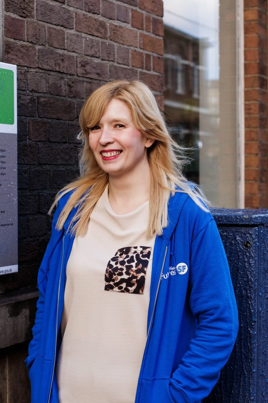 A woman with blonde hair smiling, wearing a beige shirt with leopard print detail and a blue zip-up hoodie, standing outdoors against a brick wall.