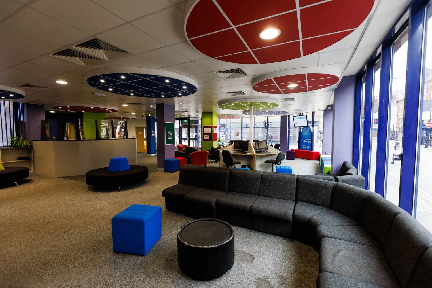 A spacious, modern lobby or waiting area with large windows, colorful circular ceiling decorations, and various seating options including a curved black sofa, blue and black ottomans, and armchairs. There are computer stations and a front desk counter.