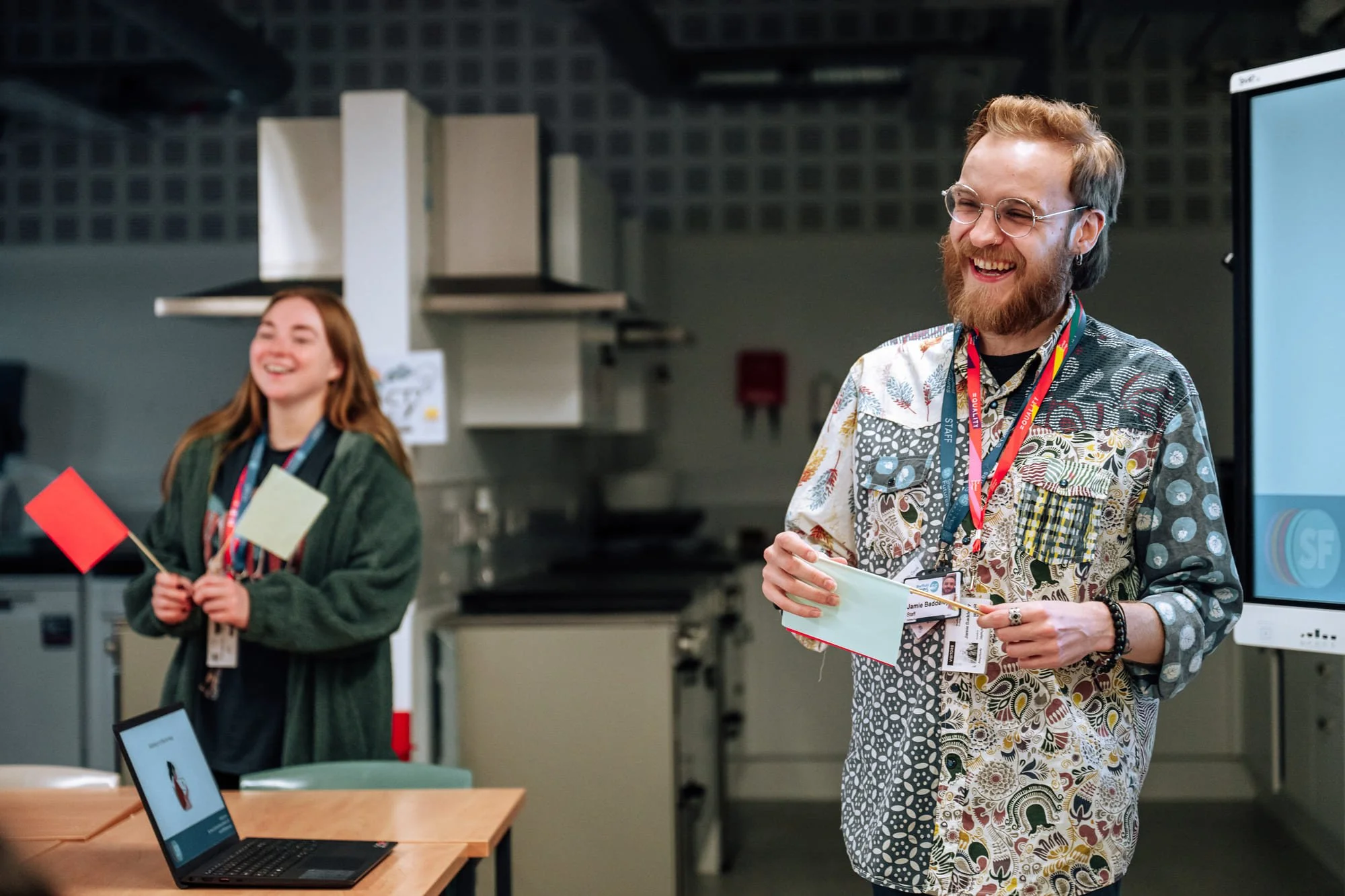 Two people smiling and presenting in a classroom or conference room. The man in front has glasses, a beard, and a colorful patterned shirt, holding a paper. The woman in the background has long hair, a green jacket, and is holding a small red flag and a paper.