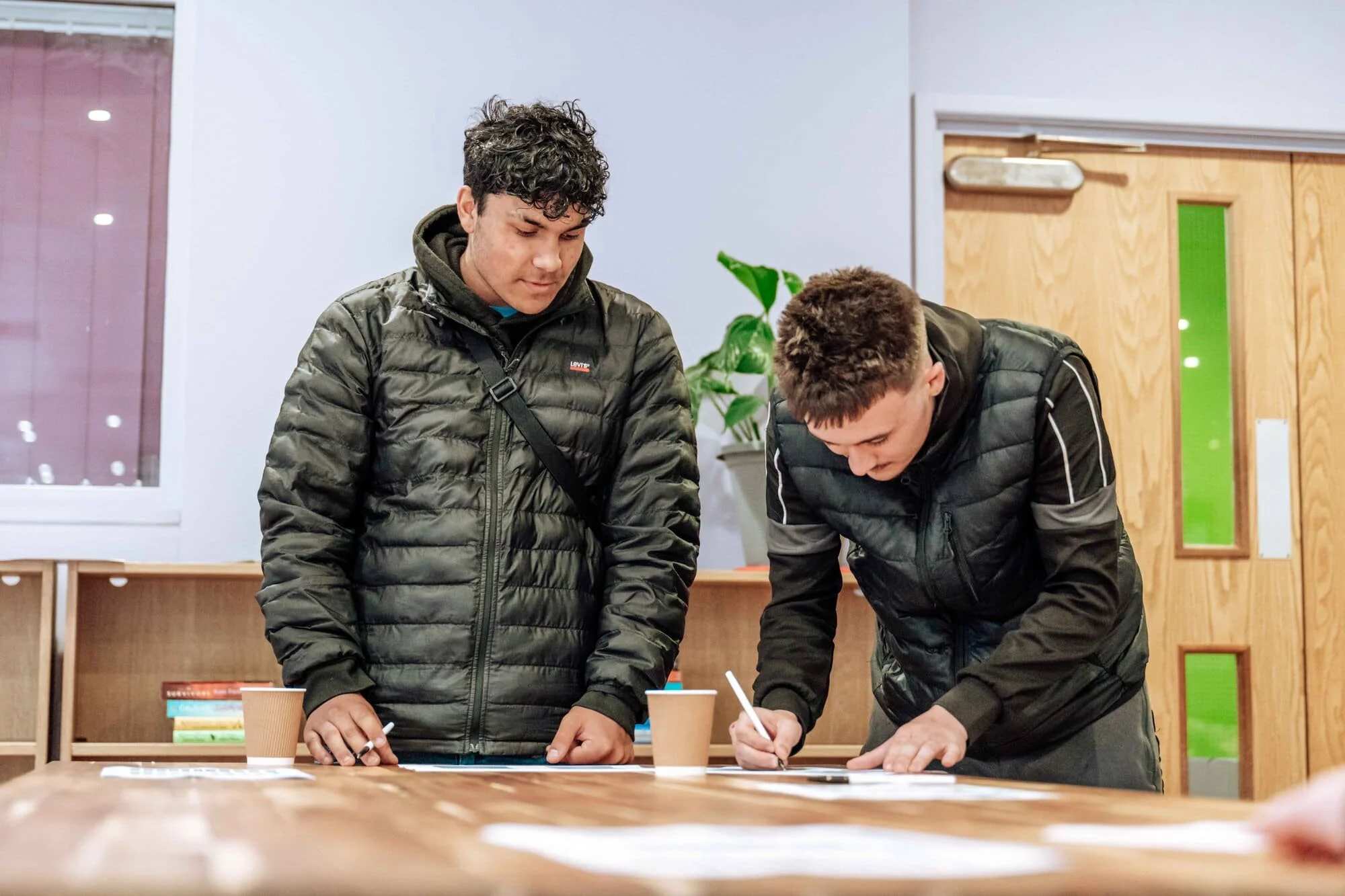 Two young men wearing black jackets are standing at a table, writing on papers in an indoor space with white walls and a wooden door.