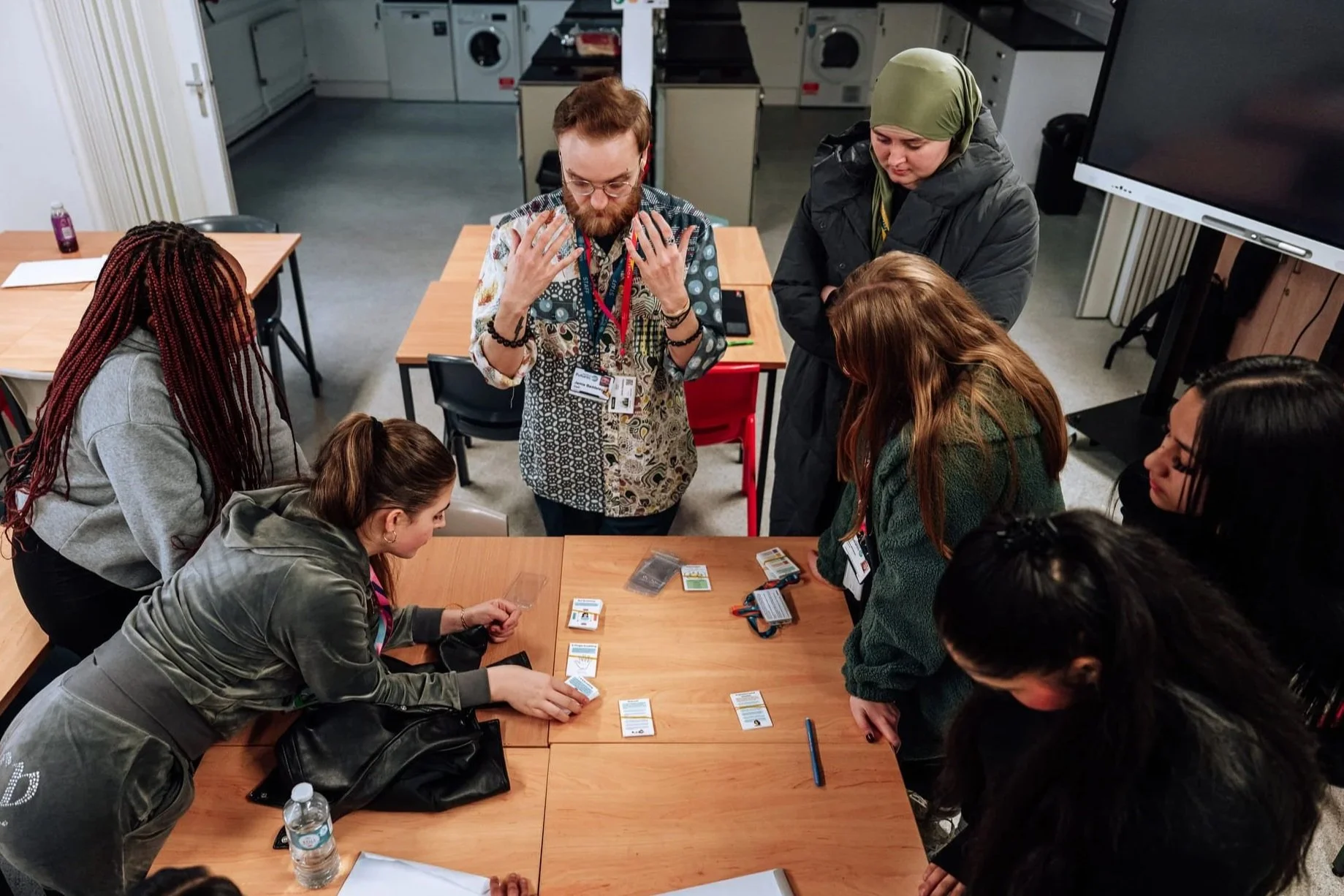 A group of college students gather around a table,  studying wellbeing cards and listening to the session leader.