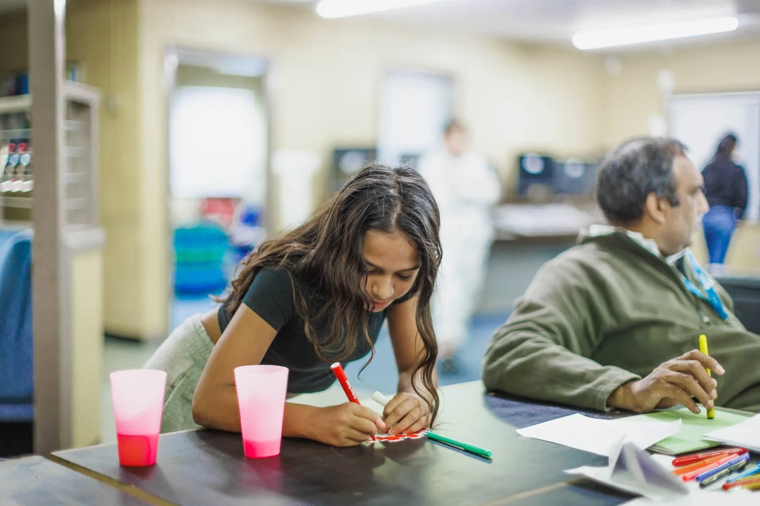 Young girl drawing with markers at a table in a classroom or art room, with two cups in front of her and a man sitting beside her, surrounded by art supplies.