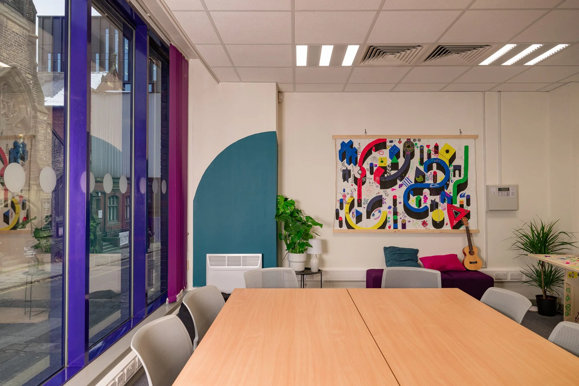A colorful modern meeting room with a wooden table, gray chairs, a purple and blue wall, a large window with purple framing, a vibrant abstract artwork on the wall, a guitar on a purple sofa, green potted plants, and a white heat register.
