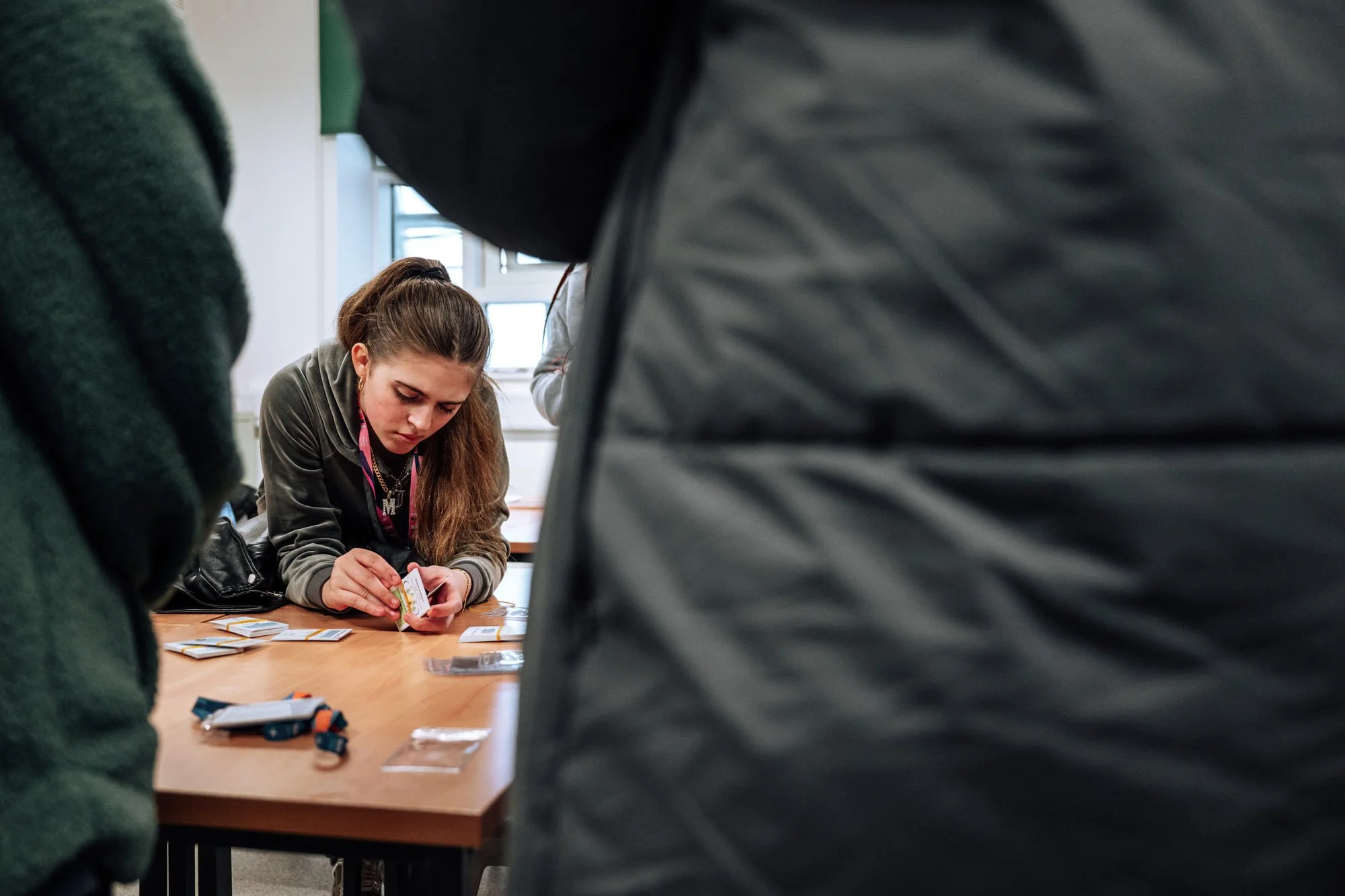 A young woman with long brown hair leaning over a wooden table, playing a card game among other people in a bright room with windows.