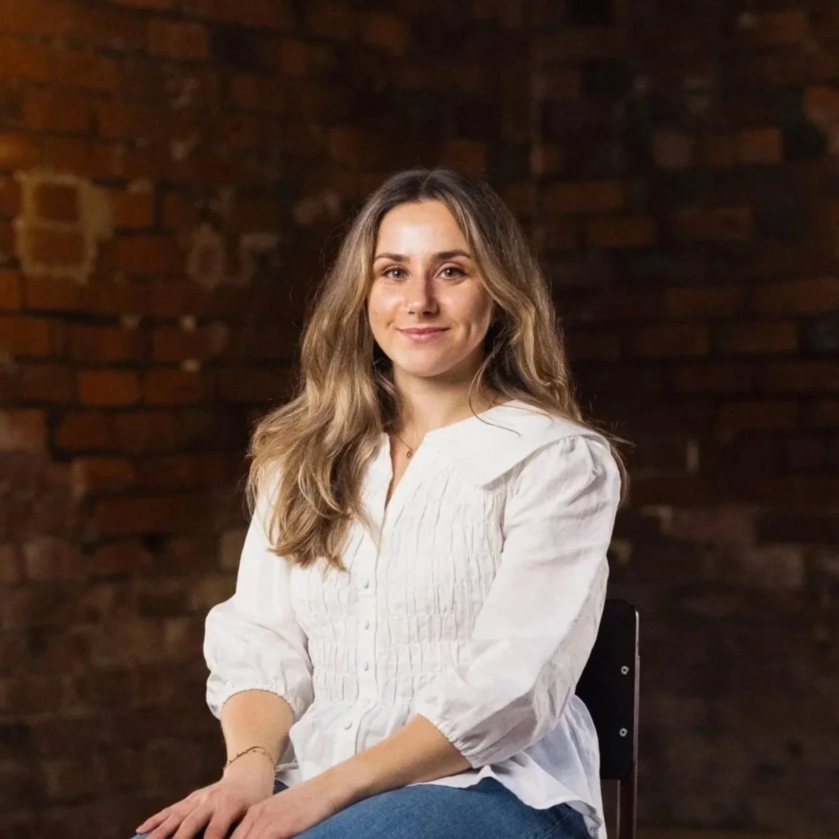 A smiling woman with shoulder-length wavy hair wearing a dark patterned blouse against a plain light gray background.