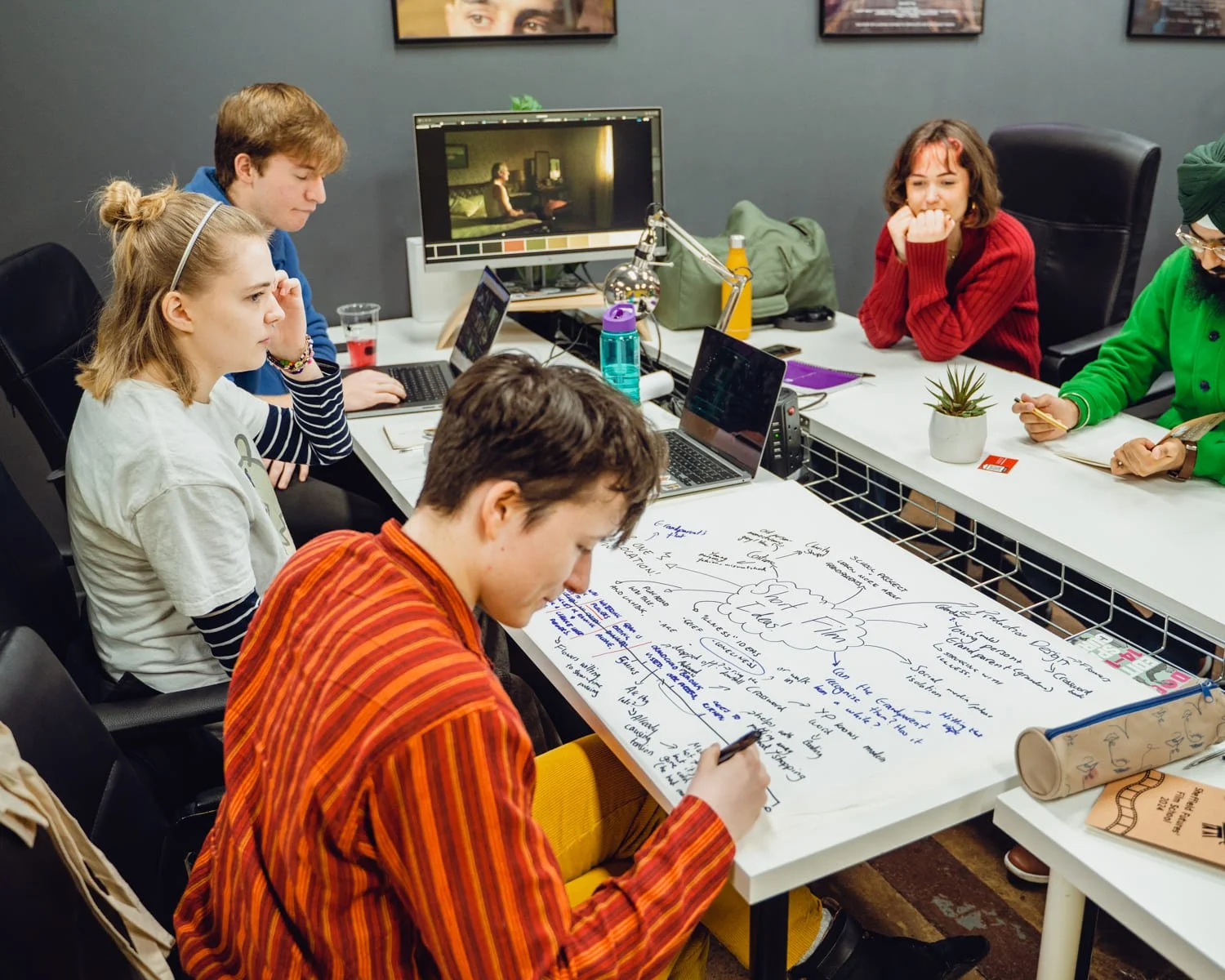 Group of young people in a meeting room, some working on laptops, others taking notes on a large paper with handwritten ideas and diagrams, surrounded by plants, books, and personal items.