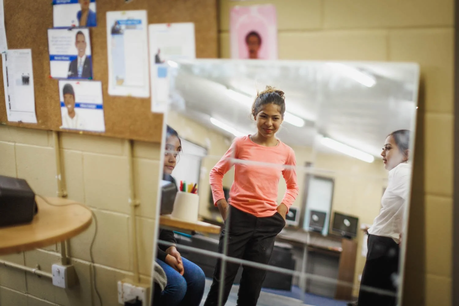 A girl in a coral shirt standing with hands in pockets, smiling, reflected in a mirror in a classroom or computer lab, with two other girls sitting and standing nearby.