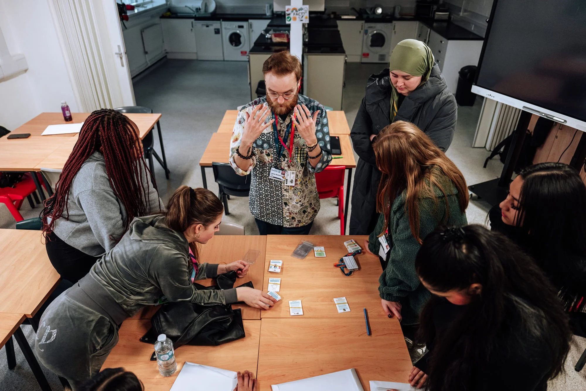 Group of people gathered around a table playing a card game, with a man explaining. There are six women and one man, and the setting appears to be a casual indoor space, possibly a community center or office meeting room.