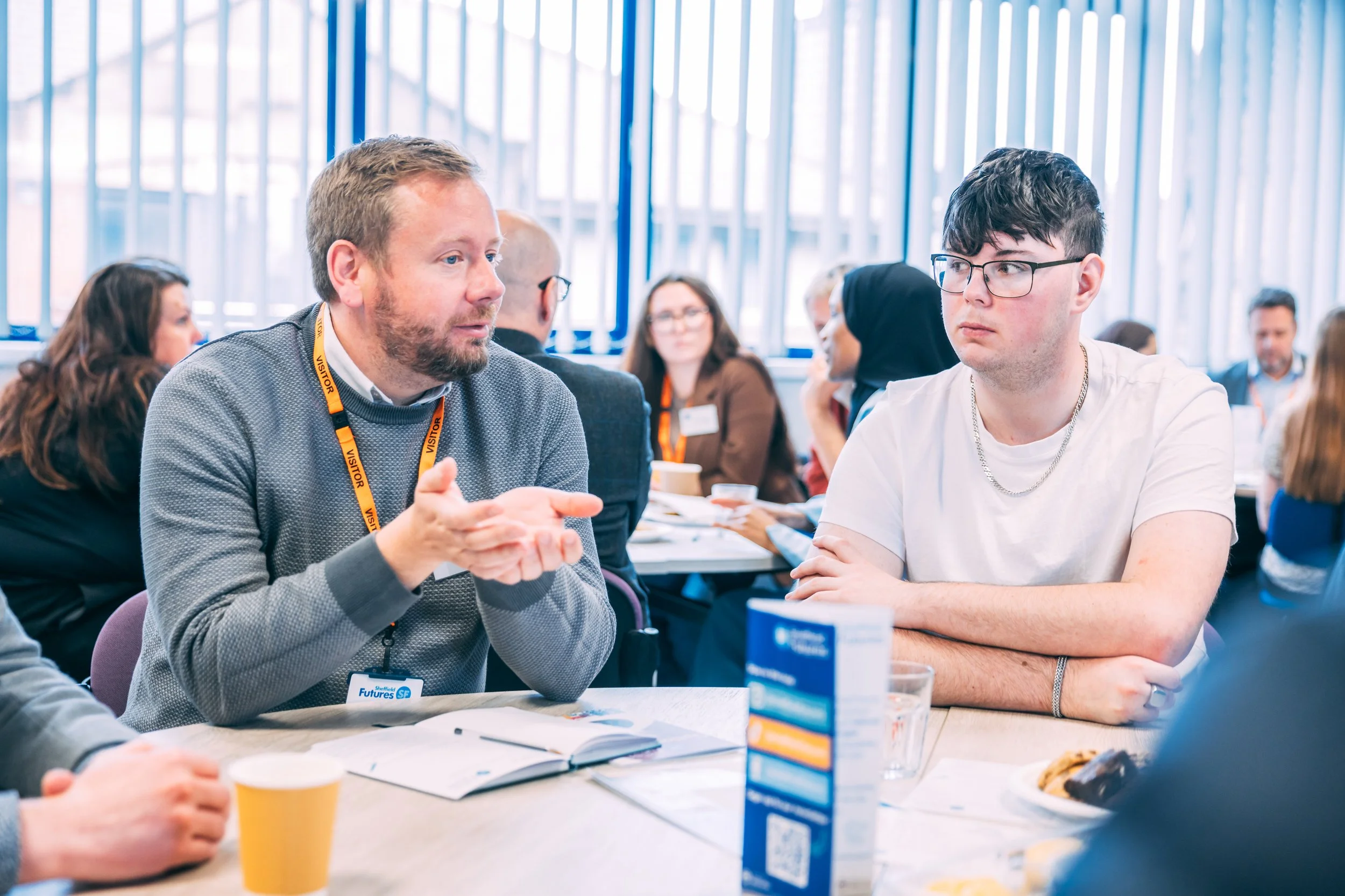 In a room full of business leaders, a young male wearing glasses sits listening to another male. In the background, groups of youth consultants are engaged in conversation with business leaders.