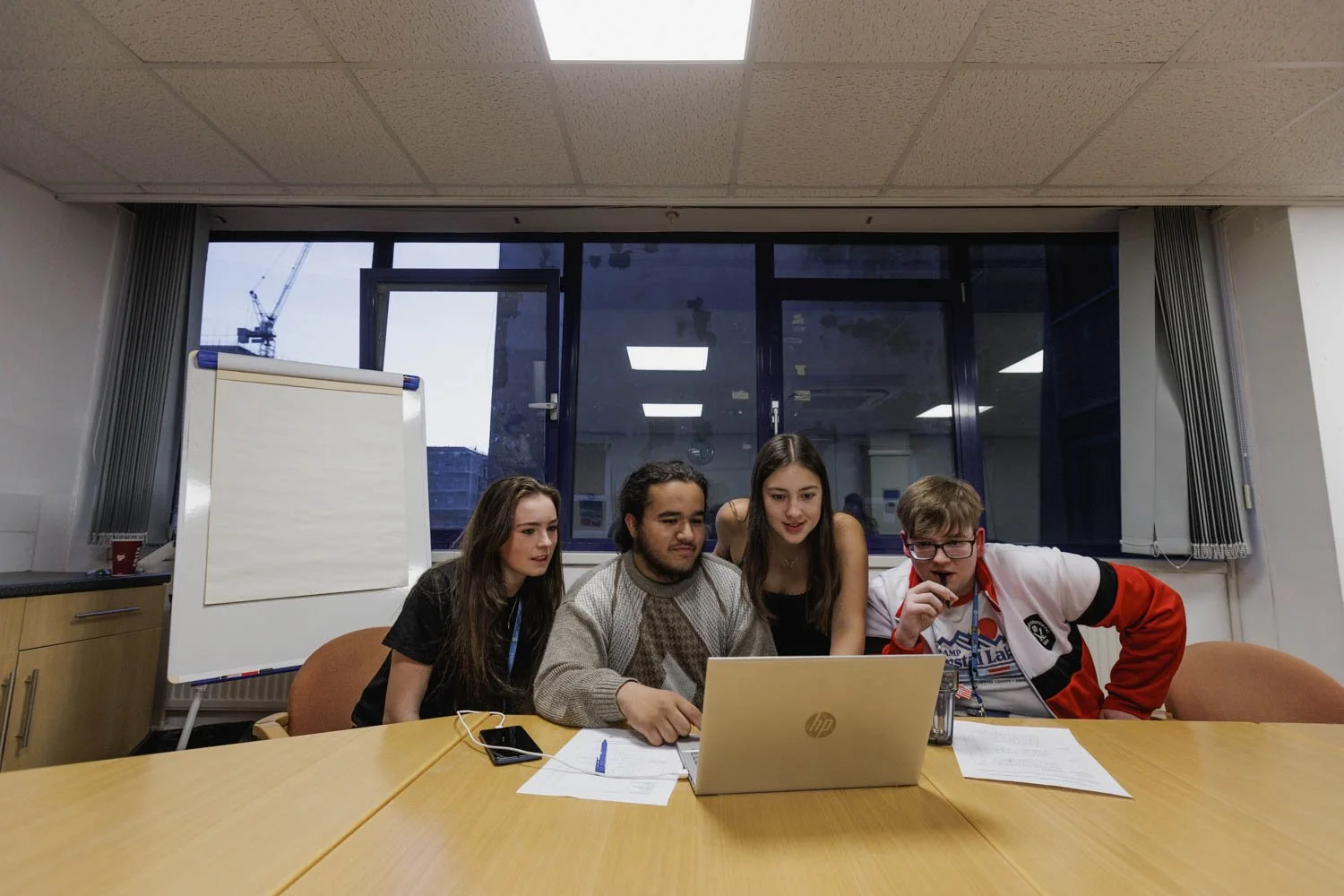 Four young adults gathered around a laptop in a conference room, looking at the screen with interest. There are documents and a smartphone on the table, and a whiteboard on a stand is visible in the background. Large windows reveal a construction crane outside.