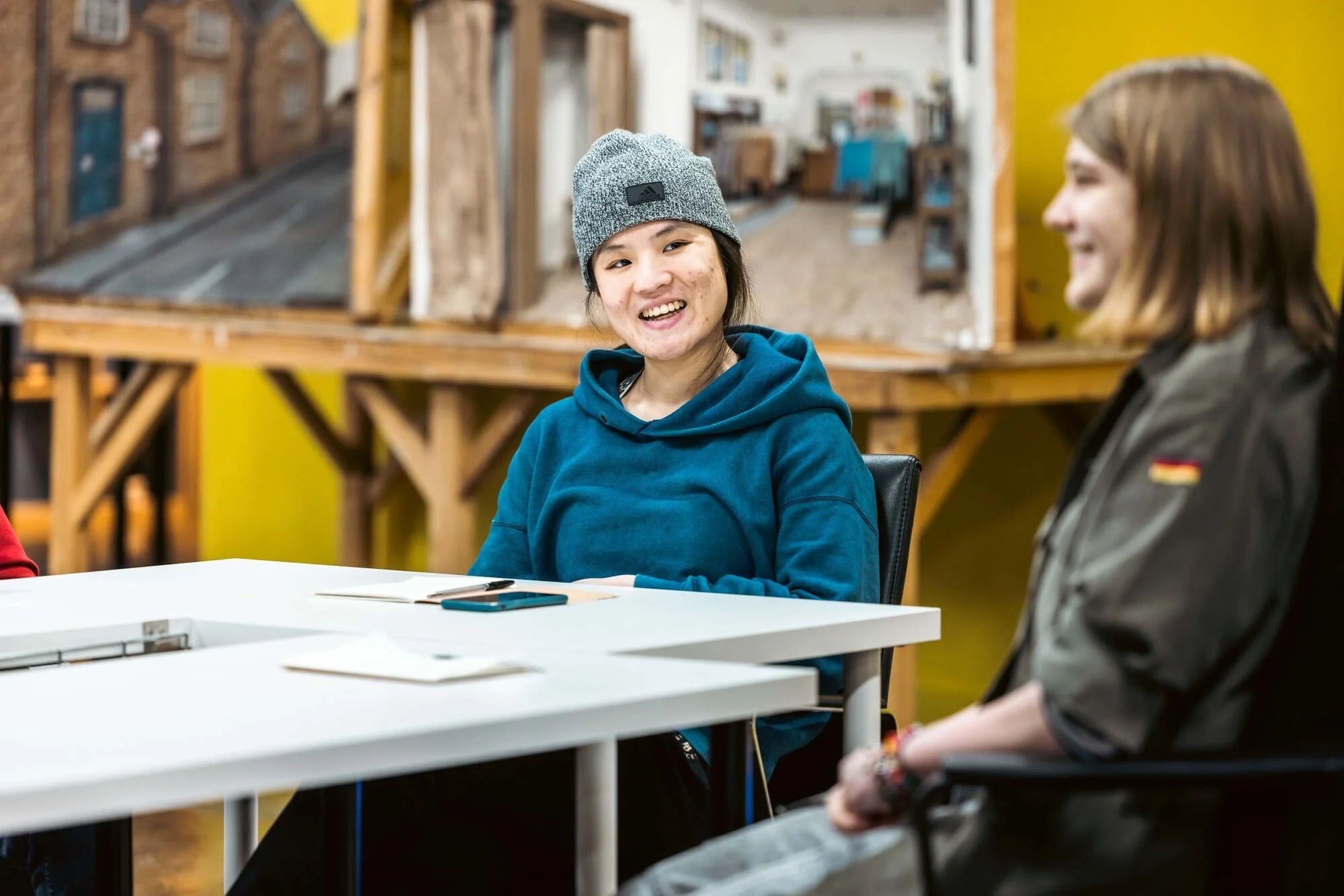 Two women sitting at a white table, smiling and talking. One woman wears a gray beanie and a blue hoodie, and the other woman has shoulder-length brown hair and wears a black jacket with a small German flag patch. The background includes a yellow wall and a model building display.