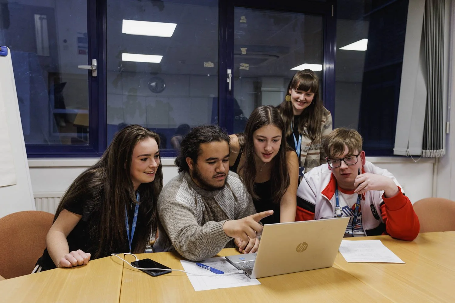 Five young people gathered around a laptop at a wooden conference table, looking at the screen and smiling, in a room with large windows and papers on the table.