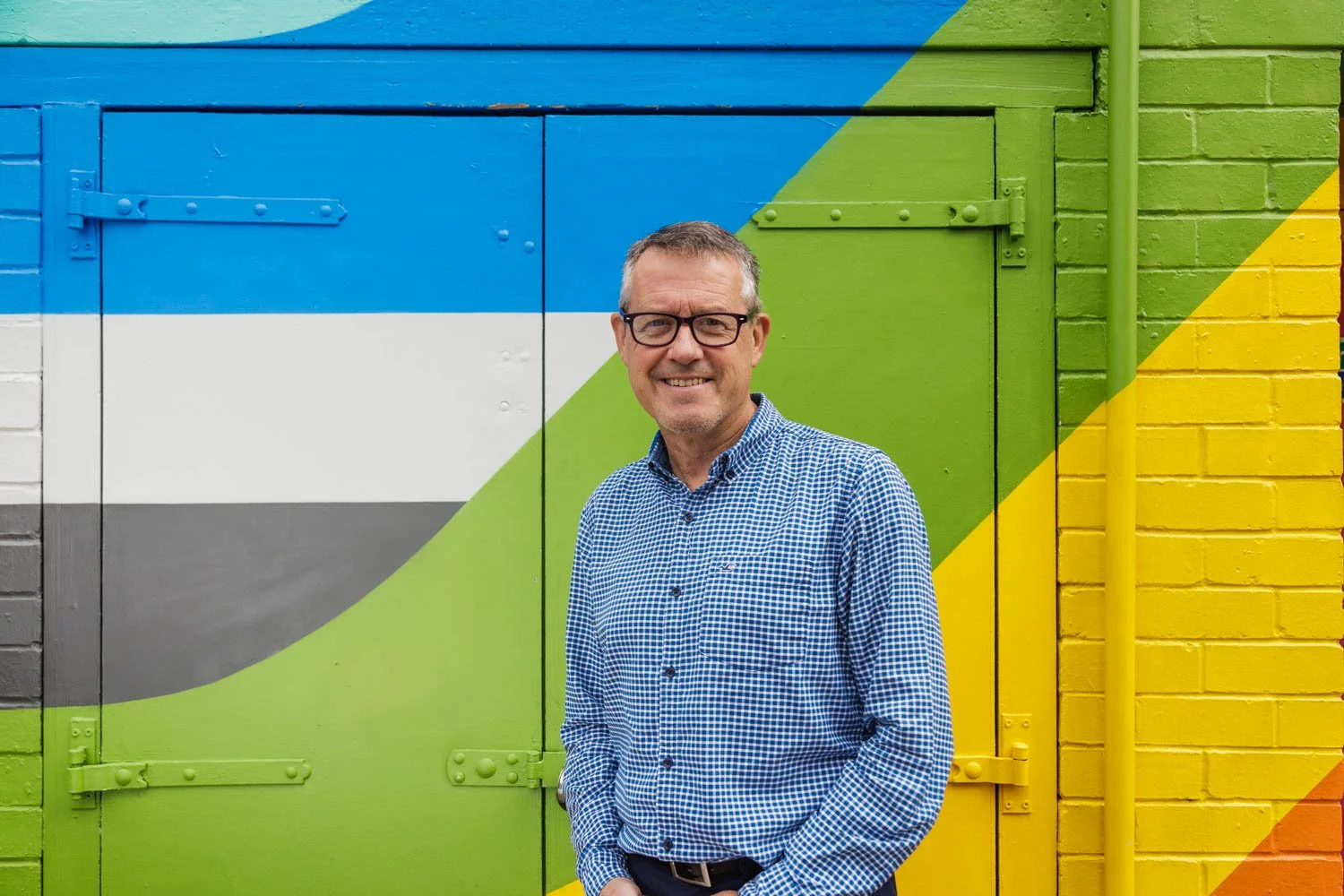 A man with glasses and a checkered blue shirt standing in front of a colorful mural with diagonal segments of blue, green, yellow, gray, and a brick wall.
