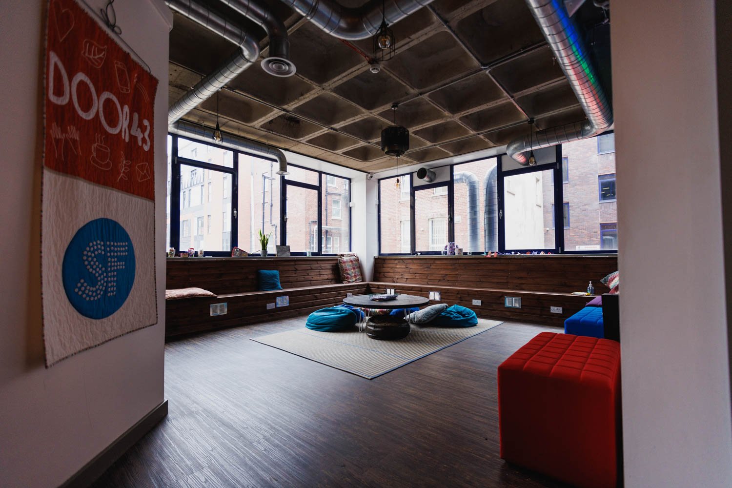 Cozy lounge with large windows, wooden benches, colorful cushions, a round coffee table, bean bags, and hanging light fixtures in an urban apartment or coworking space.