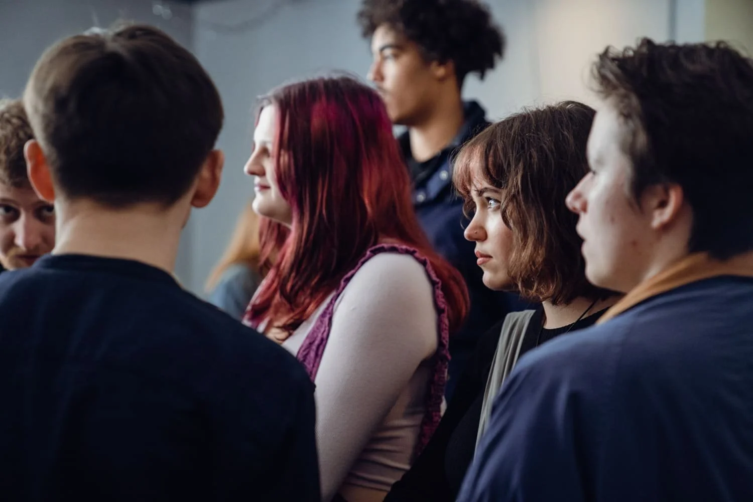 A group of young adults sitting close together, engaged in a conversation or listening to someone, in an indoor setting.