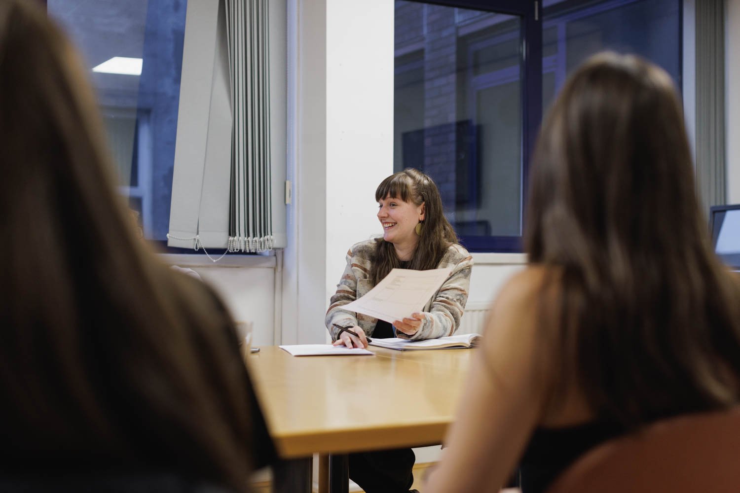 A woman woman speaking to a group of women sitting at a conference table in a meeting room, holding a paper with a bright smile.
