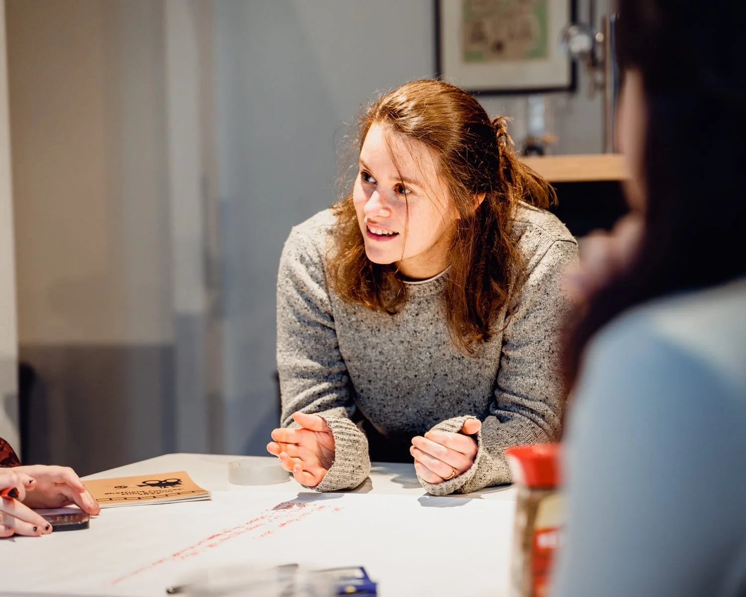 A woman with shoulder-length brown hair, wearing a gray sweater, is sitting at a table and talking to someone off-camera. There are papers, a pen, and a jar of peanut butter on the table.