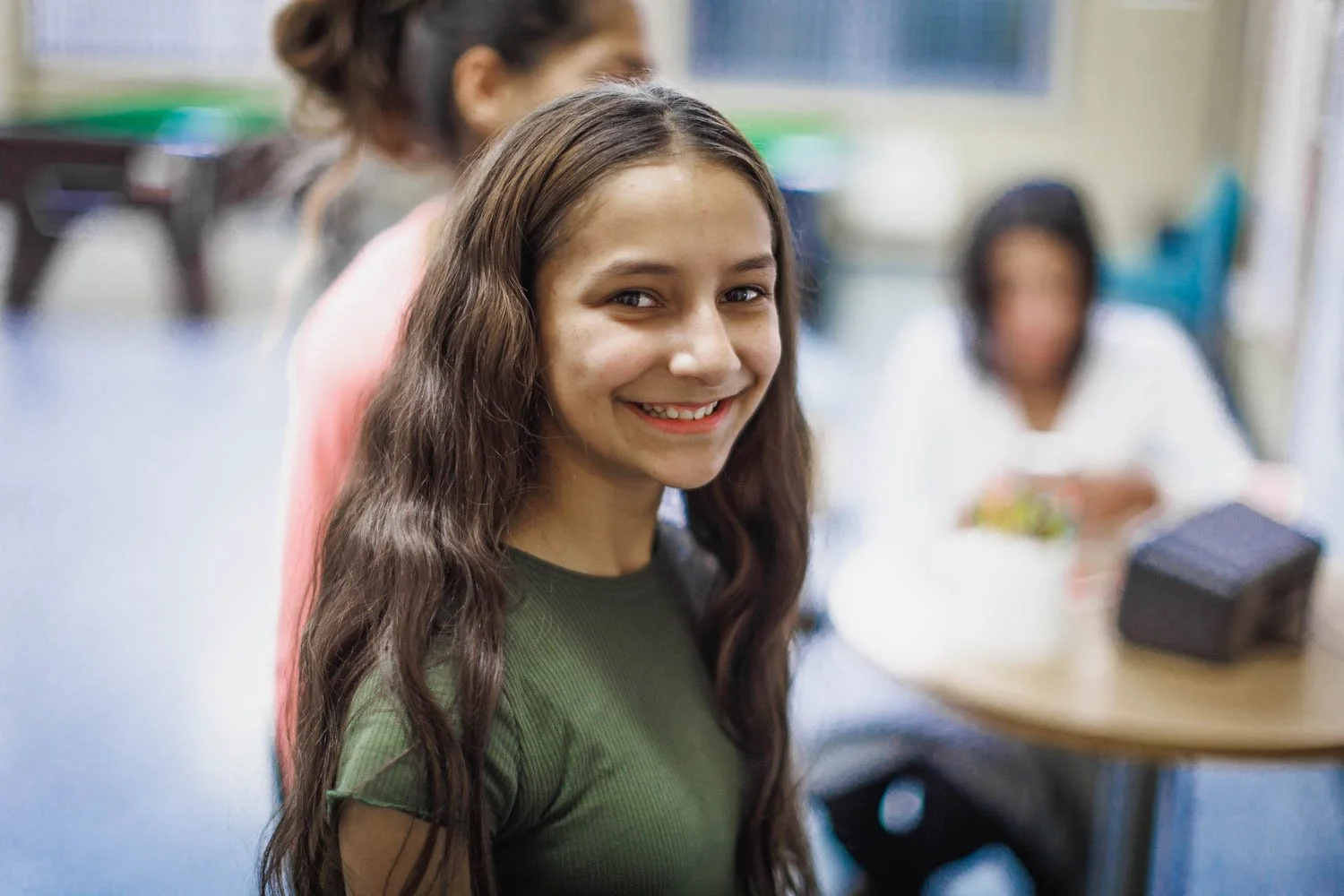 A smiling young woman with long, wavy brown hair in a classroom with other students in the background.
