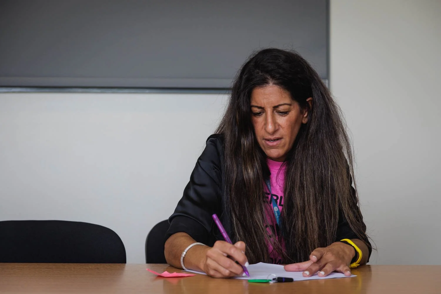 Woman with long dark hair writing with a purple pen at a desk, surrounded by colored pens, in a room with a grayboard or screen behind her.