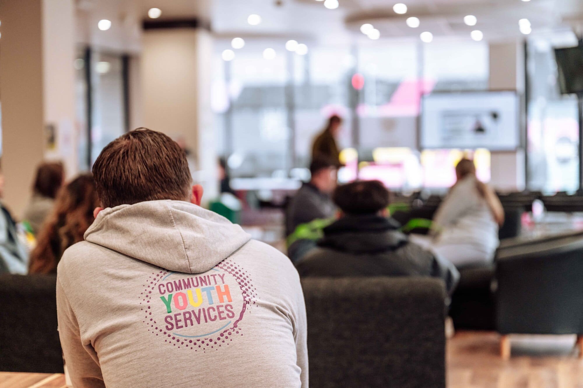 Group of young people sitting in a waiting area of a community youth services center, with one person wearing a hoodie with 'Community Youth Services' logo on the back, listening to a person speaking at the front.